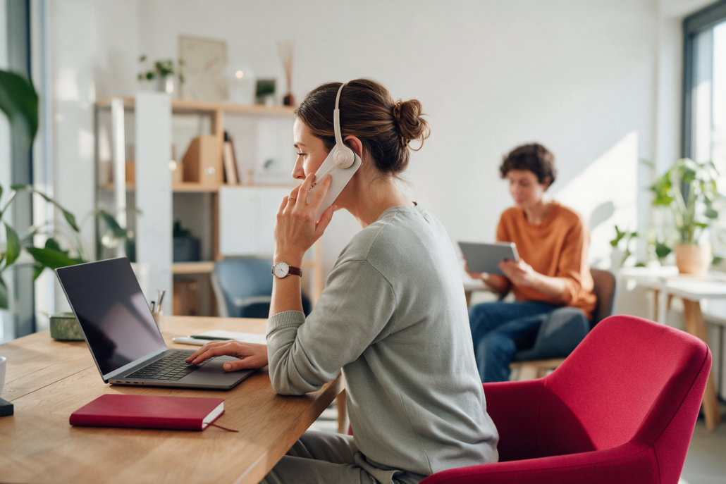 Mitarbeiterin mit Headset telefoniert am Laptop im hellen Büro, im Hintergrund eine Kollegin mit Tablet