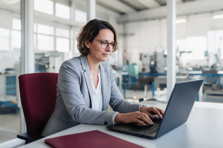 Unternehmerin arbeitet in einem hellen Büro am Laptop, im Hintergrund eine Werkhalle